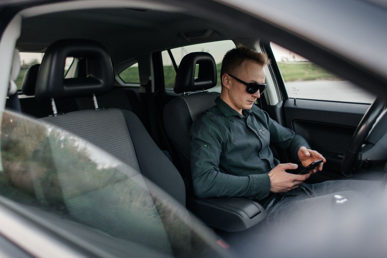 services-01 Young man using smartphone inside a parked car, wearing sunglasses.