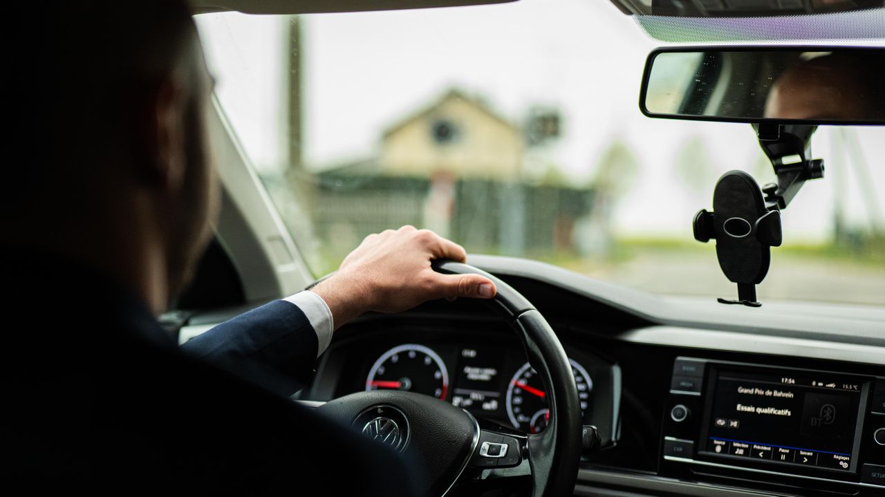 gallery-1 Businessman in suit driving a car, focus on steering wheel and dashboard.