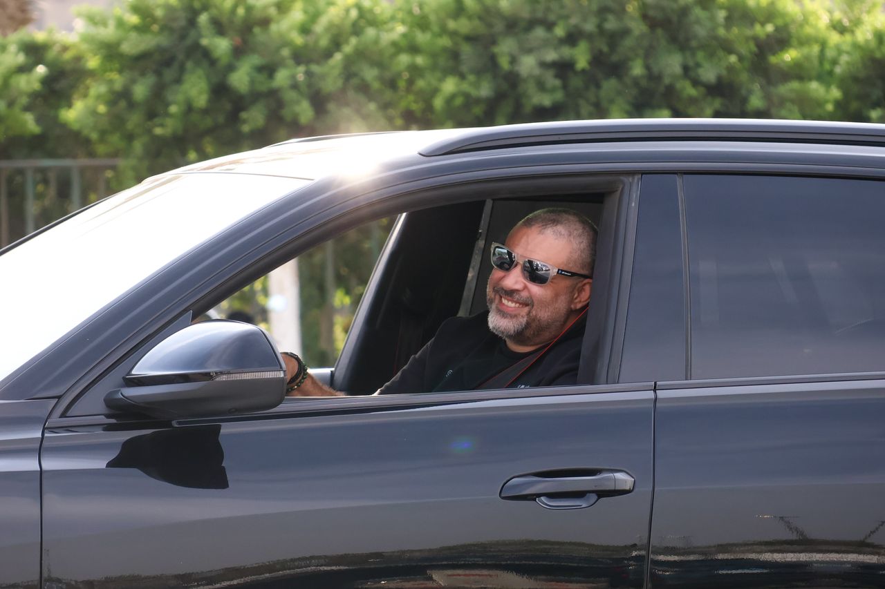 about-02 A smiling man with sunglasses driving a black car outdoors on a sunny day.