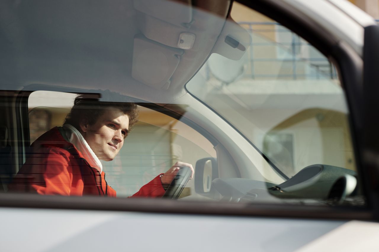 ours-journey A man in a red jacket driving a car, seen through the window on a sunny day.