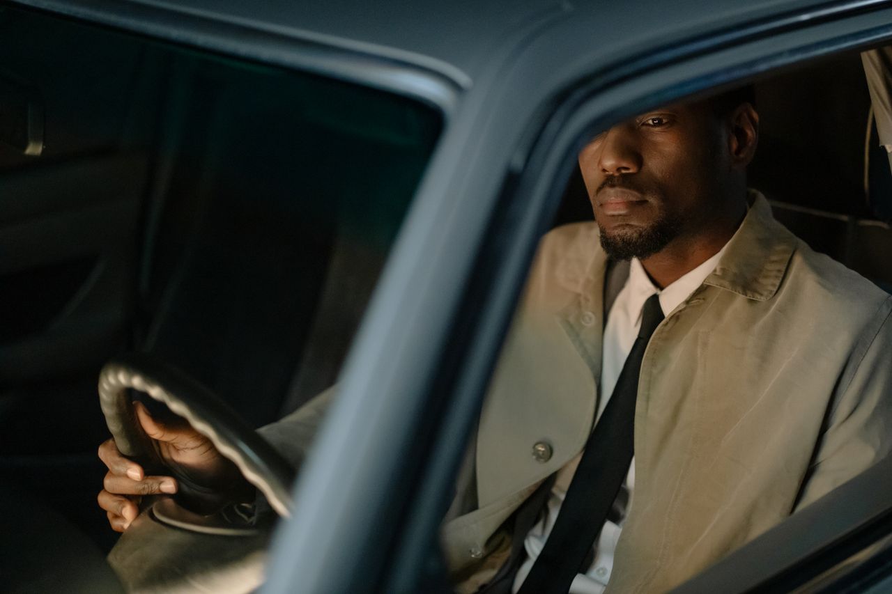 why-choose-us A man in a trench coat sits inside a car, reflecting a serious demeanor through the window.