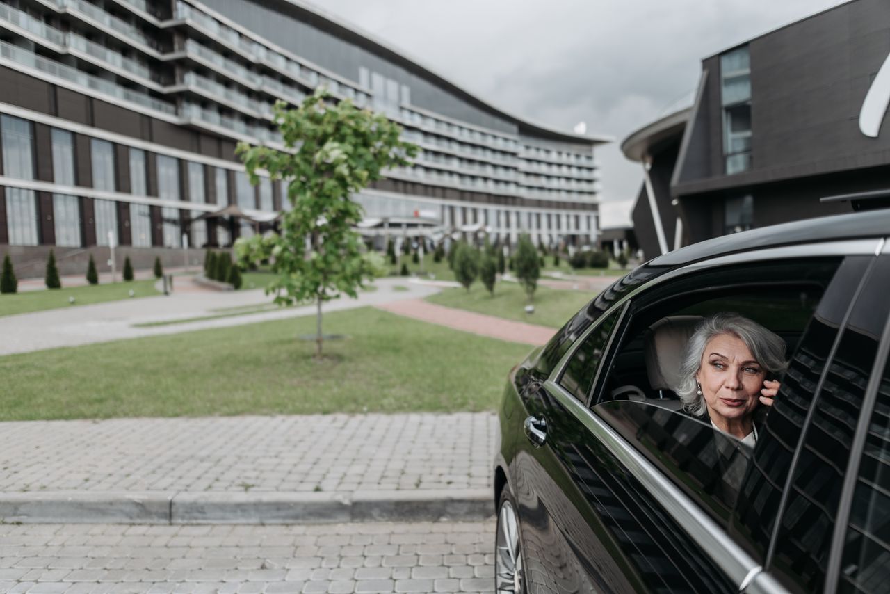 about-01 Elderly woman with grey hair on a phone call sitting in car near modern building.