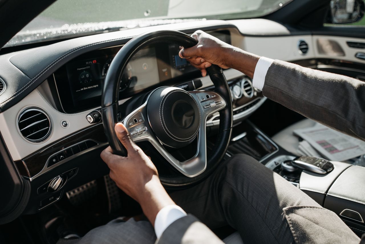 about-img Close-up of hands on steering wheel in a luxury car with dashboard and controls.