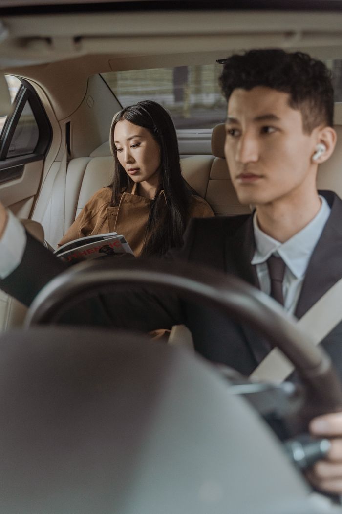 gallery-2 A businessman driving a luxury car with a passenger reading, showcasing a serene and professional travel setting.